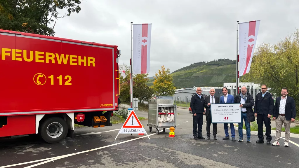 Gruppenbild bei der Spendenübergabe: Man sieht ein Feuerwehrauto, den Rollcontainer und die Gruppe bestehend aus Thomas Edringer, Leo Wächter, Matthias und Christian Krämer, sowie André Falke und Dominik Kaiser, die einen großen Spendencheck festhalten, auf dem groß „Schlauch-Rollcontainer“ steht. Im Hintergrund sieht man Fahnen und das Firmengebäude der Krämer Druck GmbH